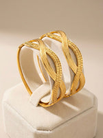A pair of micro gold-plated openable bangles featuring a textured interlocking swirl design on a cream velvet jewelry display.