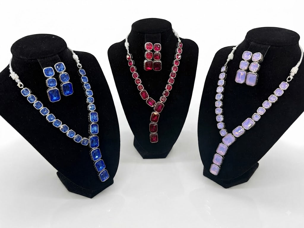 Three matching jewelry sets displayed on black velvet busts against a white background. The left set features a silver-tone necklace and earrings with rectangular blue stones.