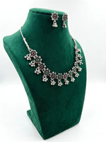 Side view of a Famaza Couture traditional oxidized silver-toned choker necklace featuring a multicolor floral lattice design with ruby and emerald stones and dangling ghungroo beads, displayed on a green velvet stand