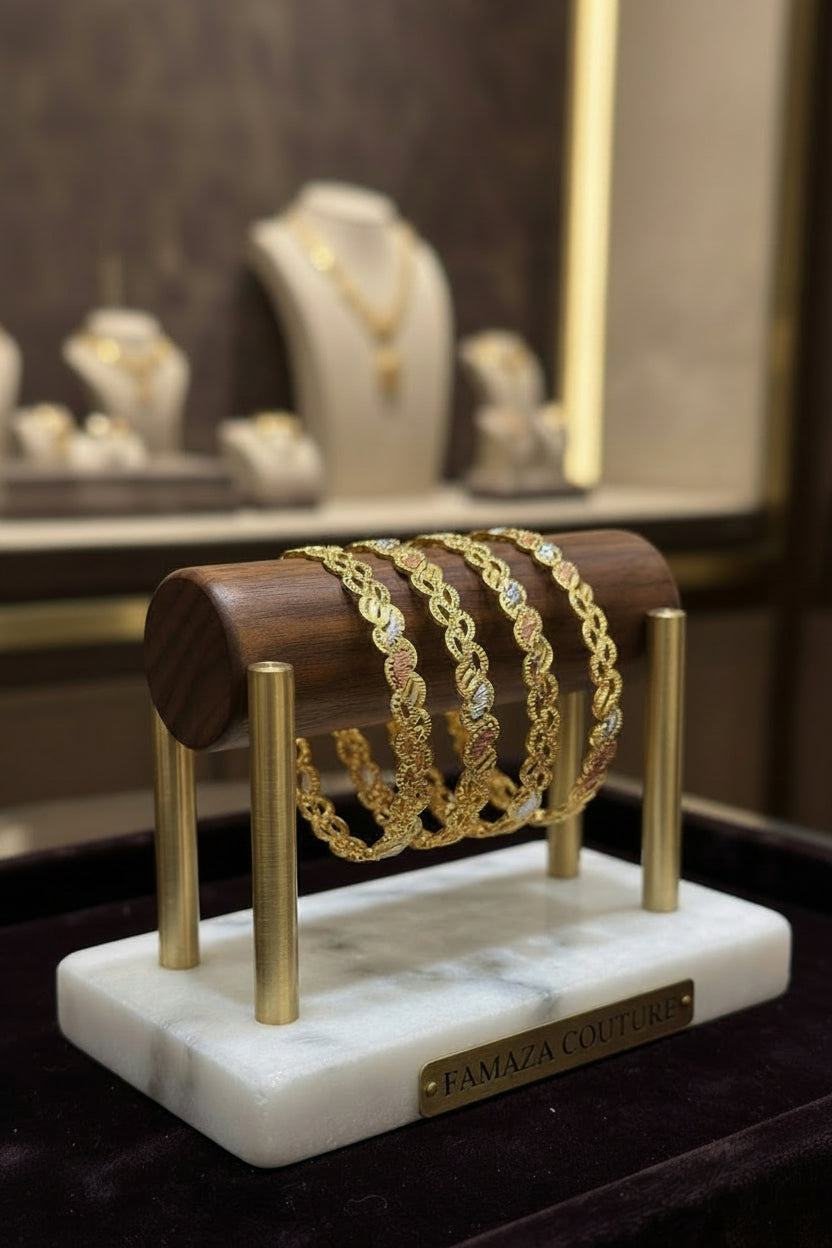 Four Famaza Couture gold, rose gold, and silver-plated interlocking wave bangles displayed on a wooden and marble stand with a metallic brand nameplate.