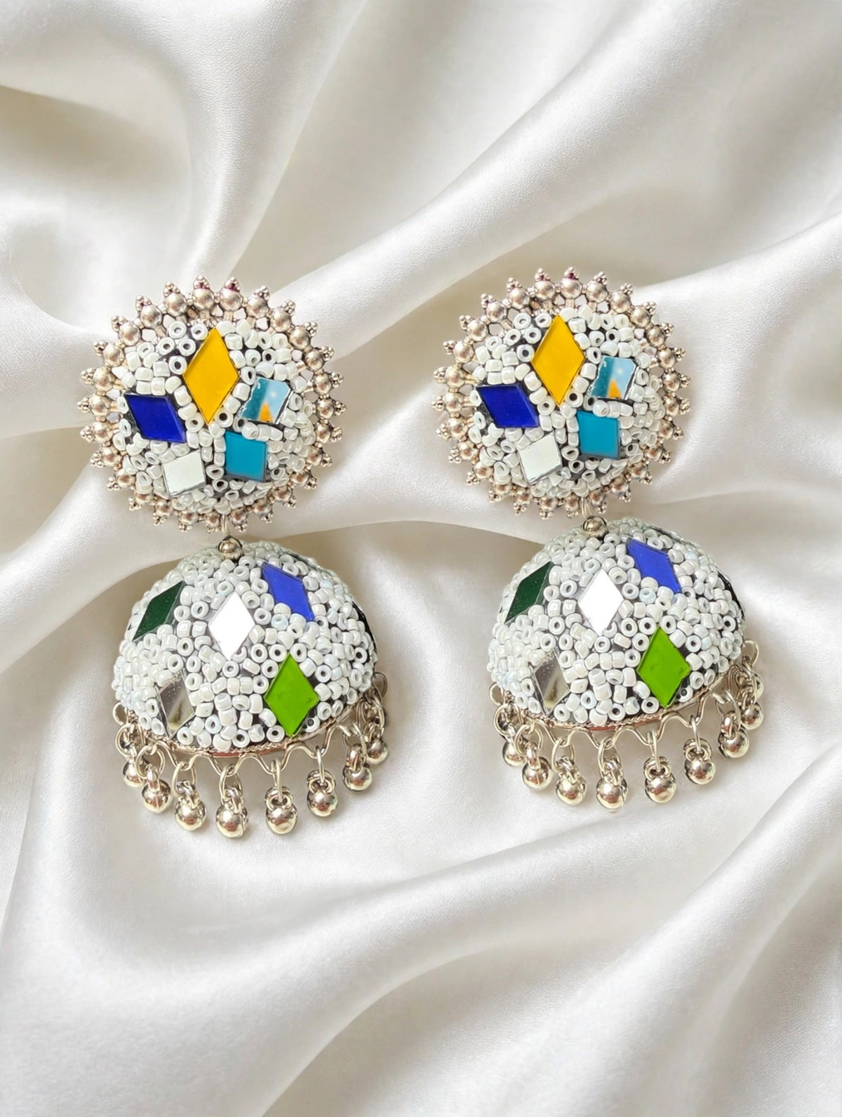 White bead multi-color meenakari jhumka — cobalt blue, golden yellow, turquoise, emerald green geometric patches, silver mirror inlays, gold-tone ghungroo drops, laid on soft draped white silk — Famaza Couture.