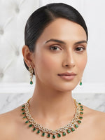 A woman with her hair pulled back wears a gold necklace and matching drop earrings featuring green teardrop-shaped stones and small clear stones, set against a light tufted wall and marble background.