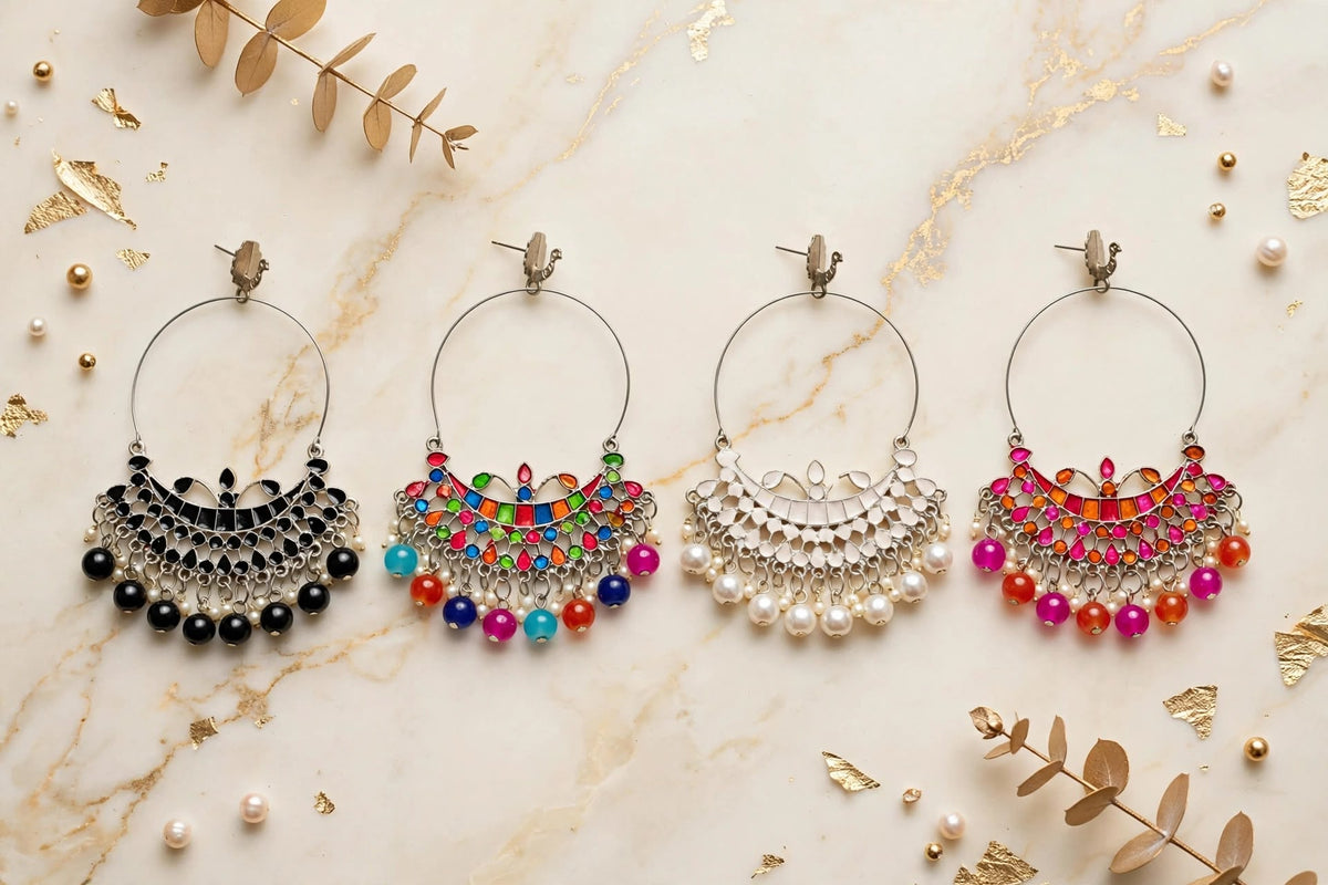 Flat-lay shot of four Chandbali silver hoop earrings arranged on cream marble with gold veining — black onyx, rainbow multi-colour, white pearl, and pink-orange Meenakari styles, accented with gold leaf and botanical sprigs.