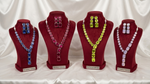 Four red velvet jewellery display busts on a white draped fabric background, each holding a matching necklace and earrings set in different gemstone colours: blue, pink, yellow-green, and light purple.