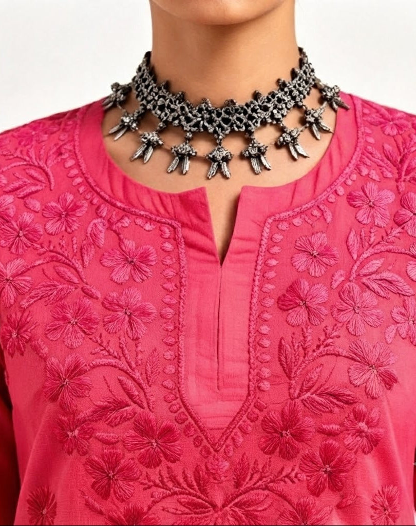 A close-up view of a person wearing a bright fuchsia pink short kurti with detailed floral embroidery along the split round neckline, styled with an ornate silver-