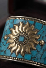 A close-up view of the Persian Royal Heirloom Turquoise Sunflower Cuff, a wide artisan bracelet featuring an intricate turquoise mosaic inlay with a prominent central gold sunflower emblem, showcased by a model in an elegant black velvet setting.