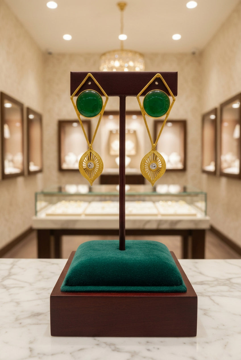 Gold-tone geometric dangle earrings with round green stones and filigree drops on a wooden stand.