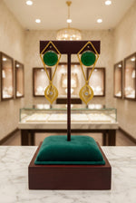 Gold-tone geometric dangle earrings with round green stones and filigree drops on a wooden stand.