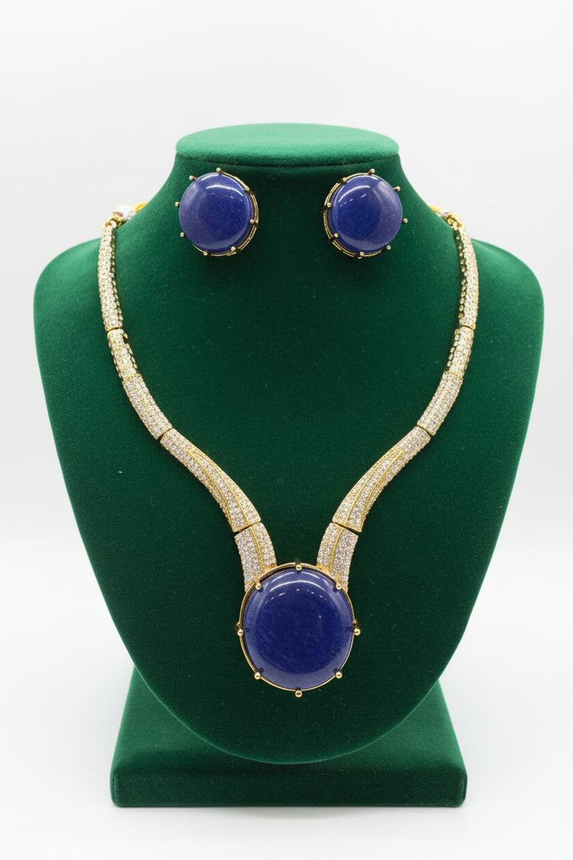 Gold-tone jewellery set featuring a necklace and matching earrings with large oval deep blue cabochon stones and rows of clear pavé stones along the chain, displayed on a white surface.