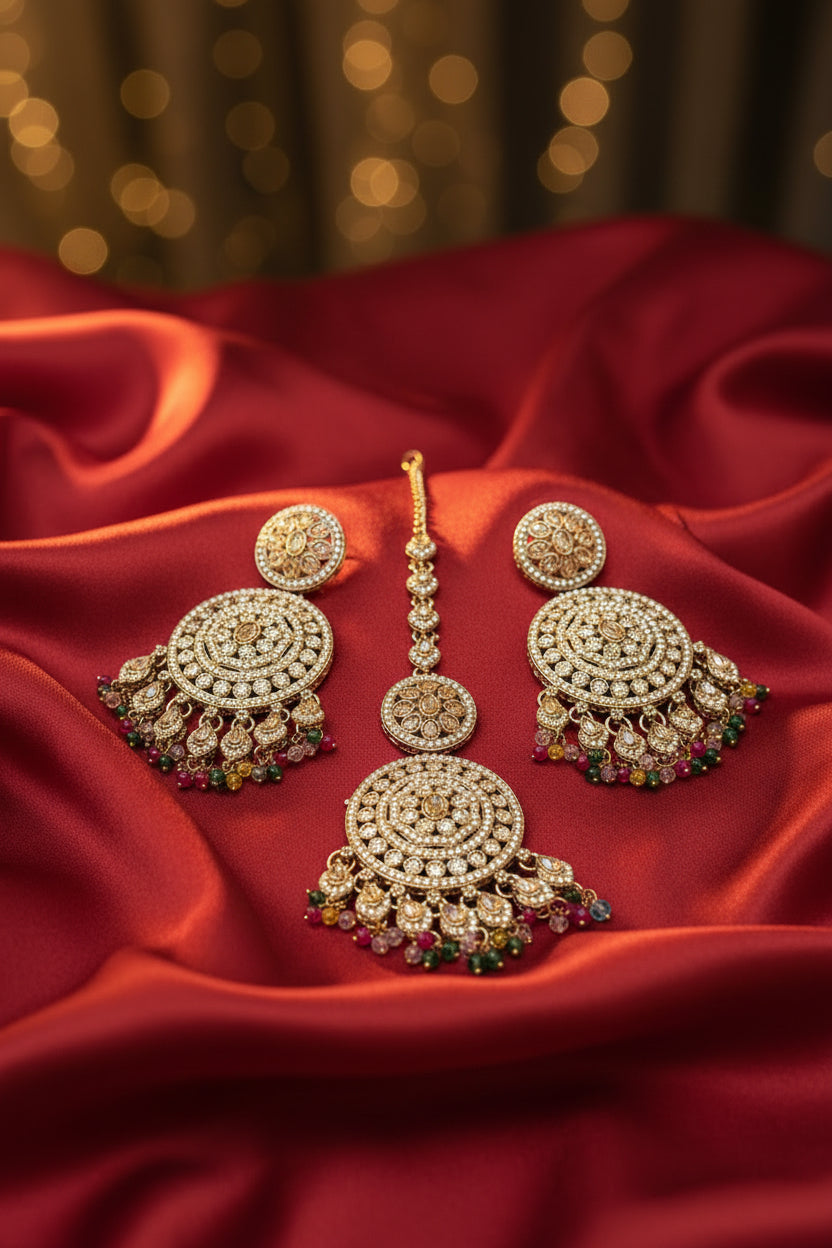 Gold-toned Indian maang tikka with matching drop earrings, decorated with clear stones and colourful pink, green, and yellow bead accents, displayed on a black textured background with a clear card labelled Famaza Couture 