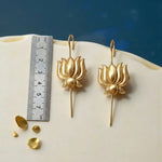 Gold lotus-shaped earrings on a white surface with a ruler for scale against a blue background.