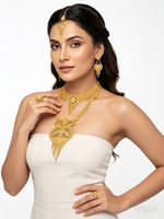 A woman modeling a gold jewelry set featuring a detailed necklace, matching earrings, and a maang tikka, paired with a white strapless dress against a plain white background.
