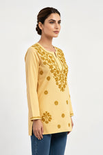 A woman modeling a light yellow long-sleeve short kurti with gold floral embroidery on the chest and scattered motifs on the front, paired with white trousers against a plain white studio background..
