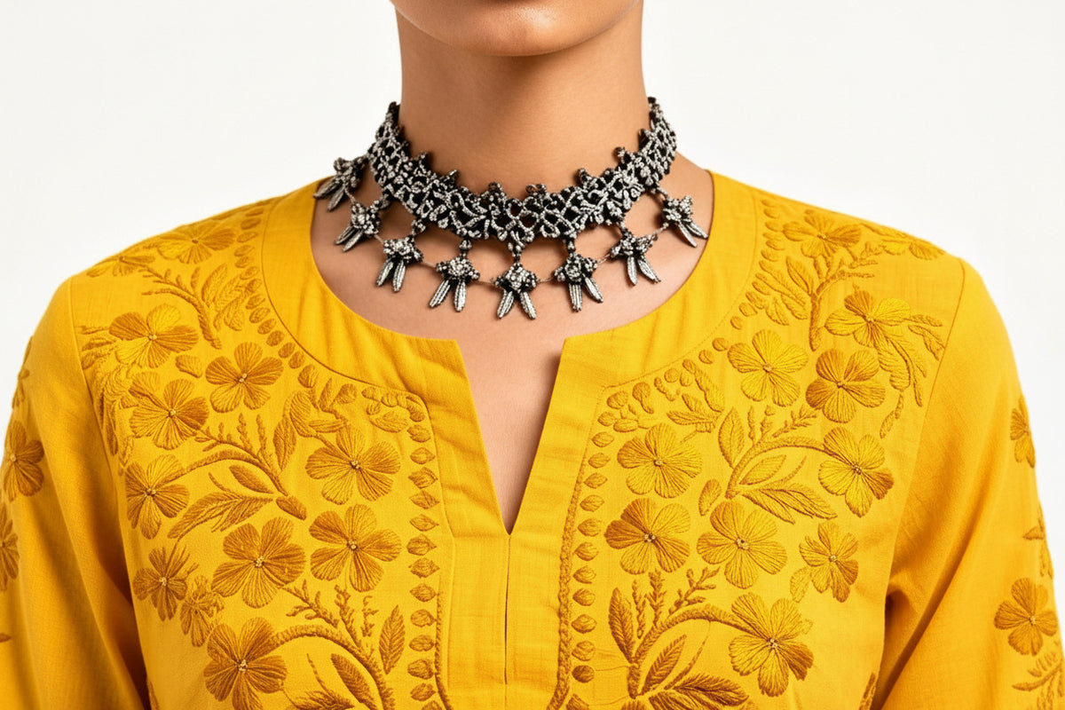 Close-up view of the neckline and chest area of a vibrant yellow cotton short kurti featuring dense, intricate floral embroidery and a keyhole neckline, styled with an ornate silver choker necklace.