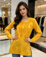 Front view of a woman wearing a vibrant yellow cotton short kurti featuring intricate matching floral embroidery, a keyhole neckline, and a square hem line, paired with blue jeans and posing with her hands on her hips in an indoor boutique setting.