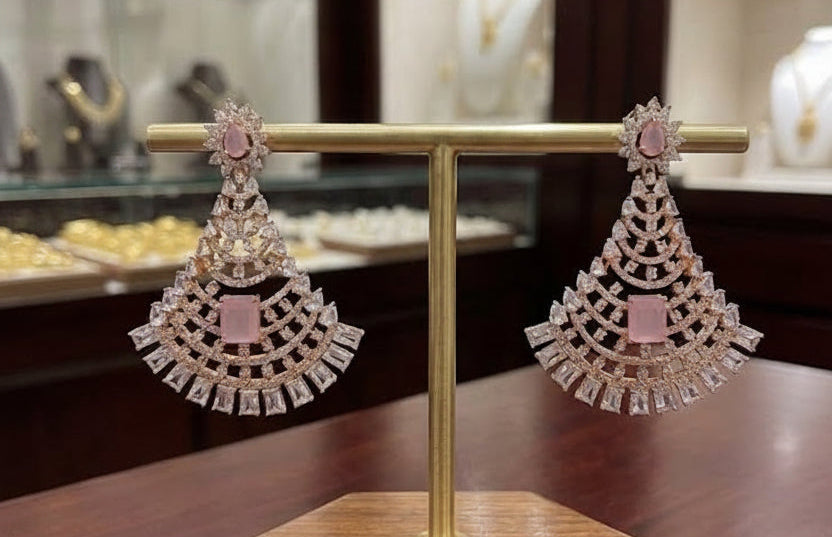A pair of ornate dangling earrings displayed on a gold T-stand with a wooden base in a jewelry store. The earrings feature pink stones and clear rhinestones, with a blurred background of jewelry displays.