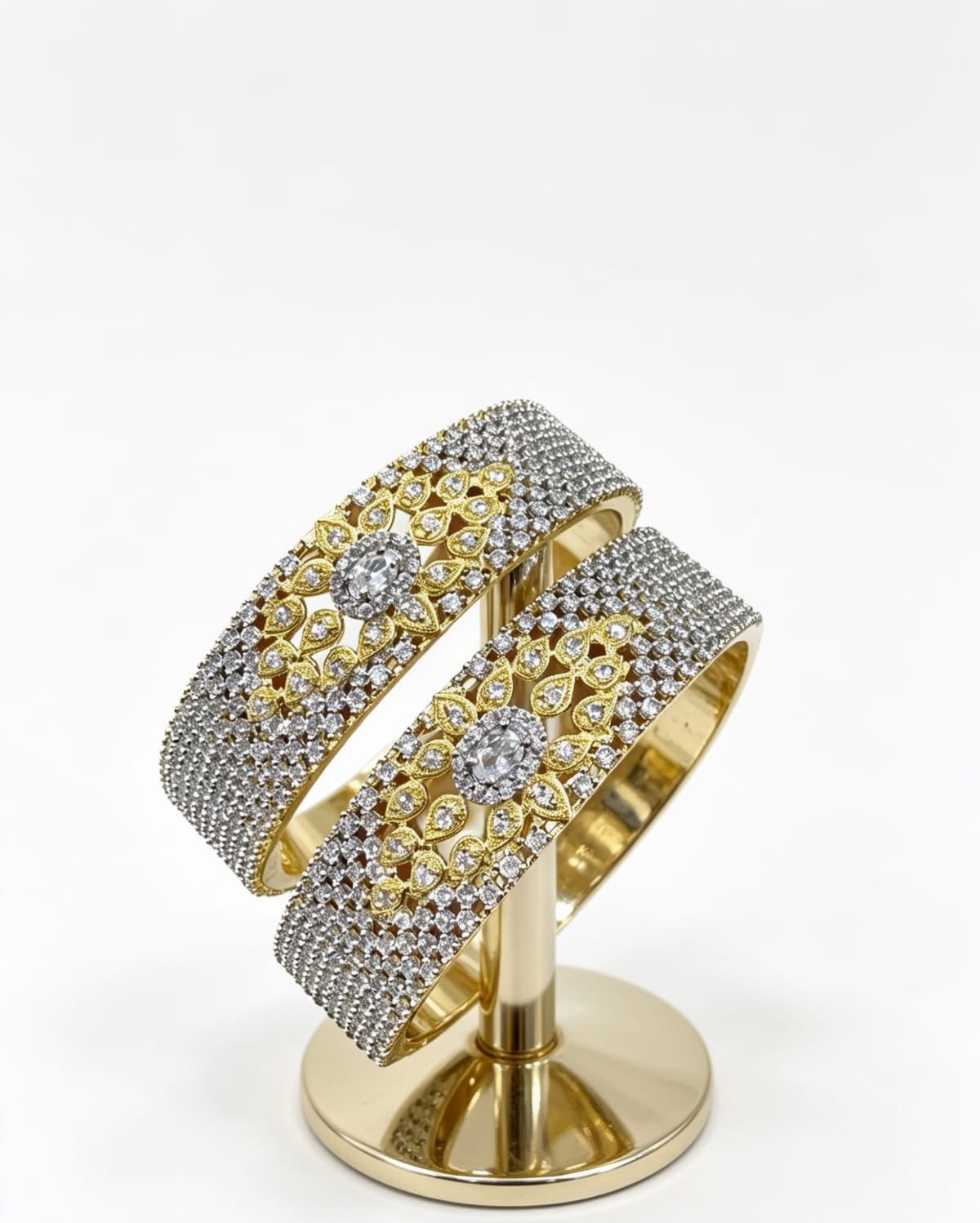 A pair of wide-band dual-tone American Diamond bangles with a central gold floral design, displayed on a professional gold jewelry stand.