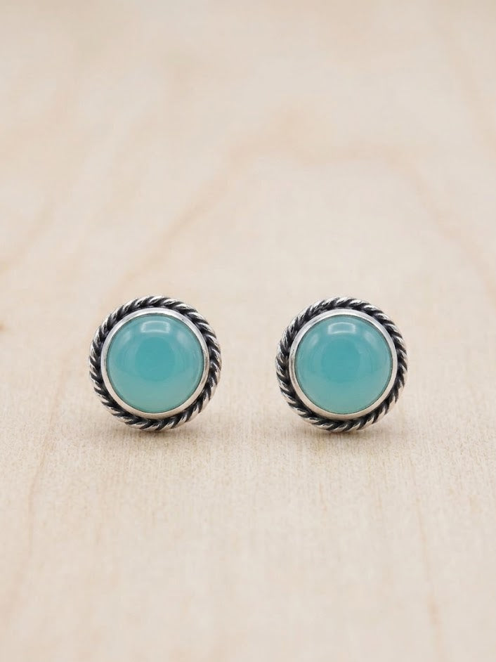 A pair of round stud earrings with smooth light blue-green cabochon stones in a silver-toned twisted rope bezel setting, displayed on a light wood surface.