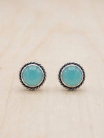 A pair of round stud earrings with smooth light blue-green cabochon stones in a silver-toned twisted rope bezel setting, displayed on a light wood surface.