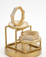 Close-up of a luxury hexagonal gold-plated bangle featuring intricate pavé-set crystals and prominent baguette-cut American diamonds on a tiered gold display stand against a white background.