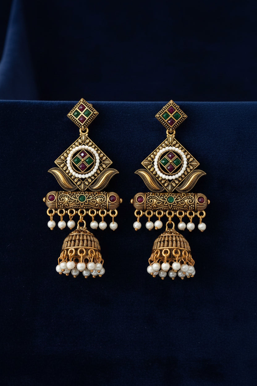 A hand holding a pair of ornate Indian gold-plated earrings with green and red enamel, white pearl borders, and dangling pearl drops, set against a plain off-white background.