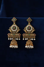 A hand holding a pair of ornate Indian gold-plated earrings with green and red enamel, white pearl borders, and dangling pearl drops, set against a plain off-white background.