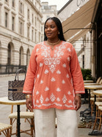 A woman stands outdoors in a city street setting wearing a peach-orange tunic top with white floral embroidery and white pants. A patterned black and beige handbag with a visible Christian Dior logo sits on a table beside her.