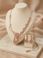 Rose gold jewelry set displayed on a cream jewelry bust, including a necklace with floral motifs, matching earrings, and a chain with a small box, set against a warm beige satin background.