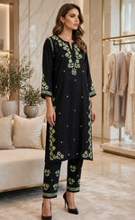 A woman models a black three-piece kurta set with green floral embroidery in a modern boutique setting.