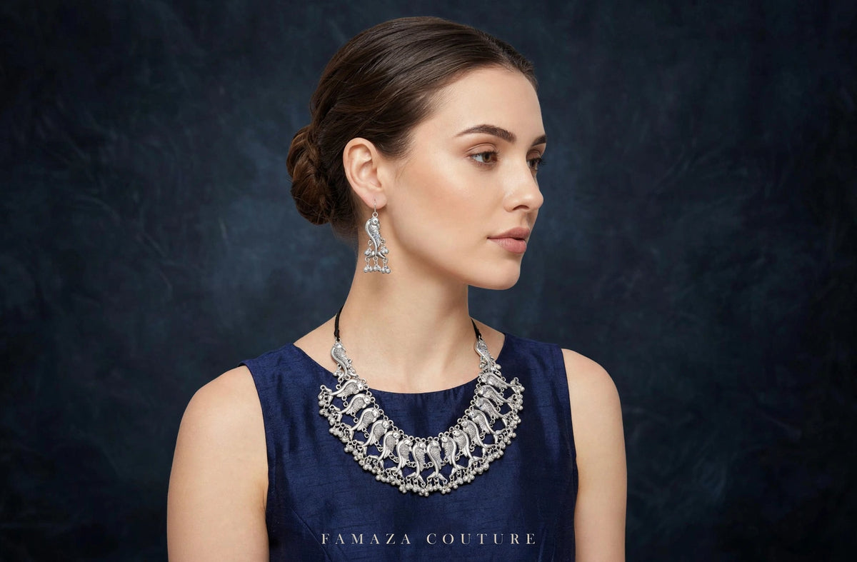 A woman in profile wearing a dark navy sleeveless top and a large silver statement necklace with matching drop earrings, set against a dark textured studio background. The brand name 'FAMAZA COUTURE' is printed at the bottom of the image.