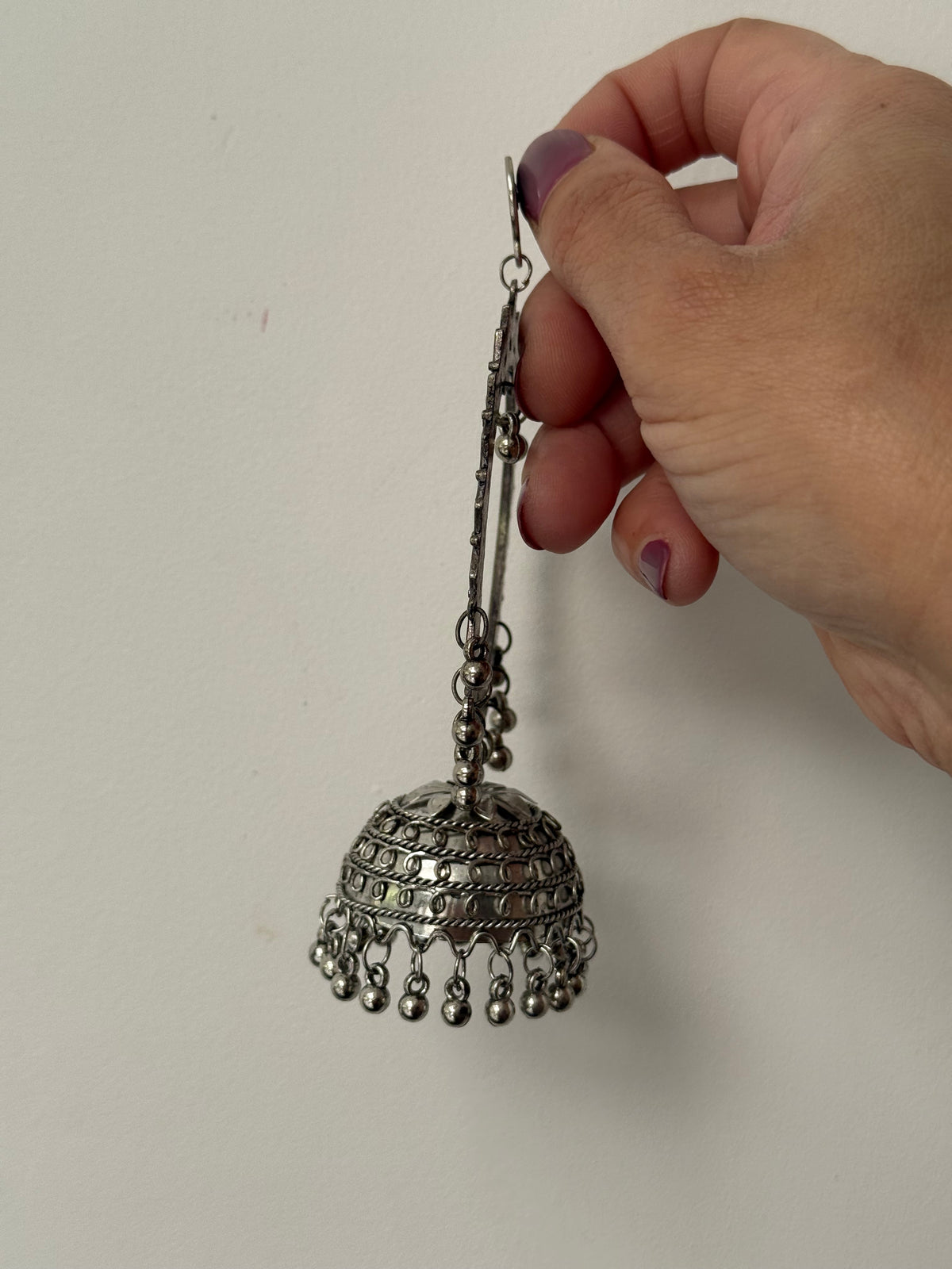 A hand with purple nail polish holds a silver-toned, ornate jhumka earring against a plain off-white background.
