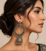 A hand holding a large silver-toned traditional hoop earring decorated with blue square stones and dangling bell-shaped charms, shown against a plain off-white background. Total drop length is 14cms and hoop width is 7cms the bottom dome width is 5cms 