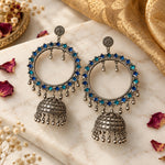 A pair of large silver-toned hoop earrings with blue rhinestone accents and dangling jhumka bells, held in a person's hand against a plain light background.