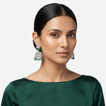 A woman with dark hair pulled back wears a pair of ornate silver-toned jhumka earrings with green stone accents. She is wearing a dark green satin top against a light gray studio background.