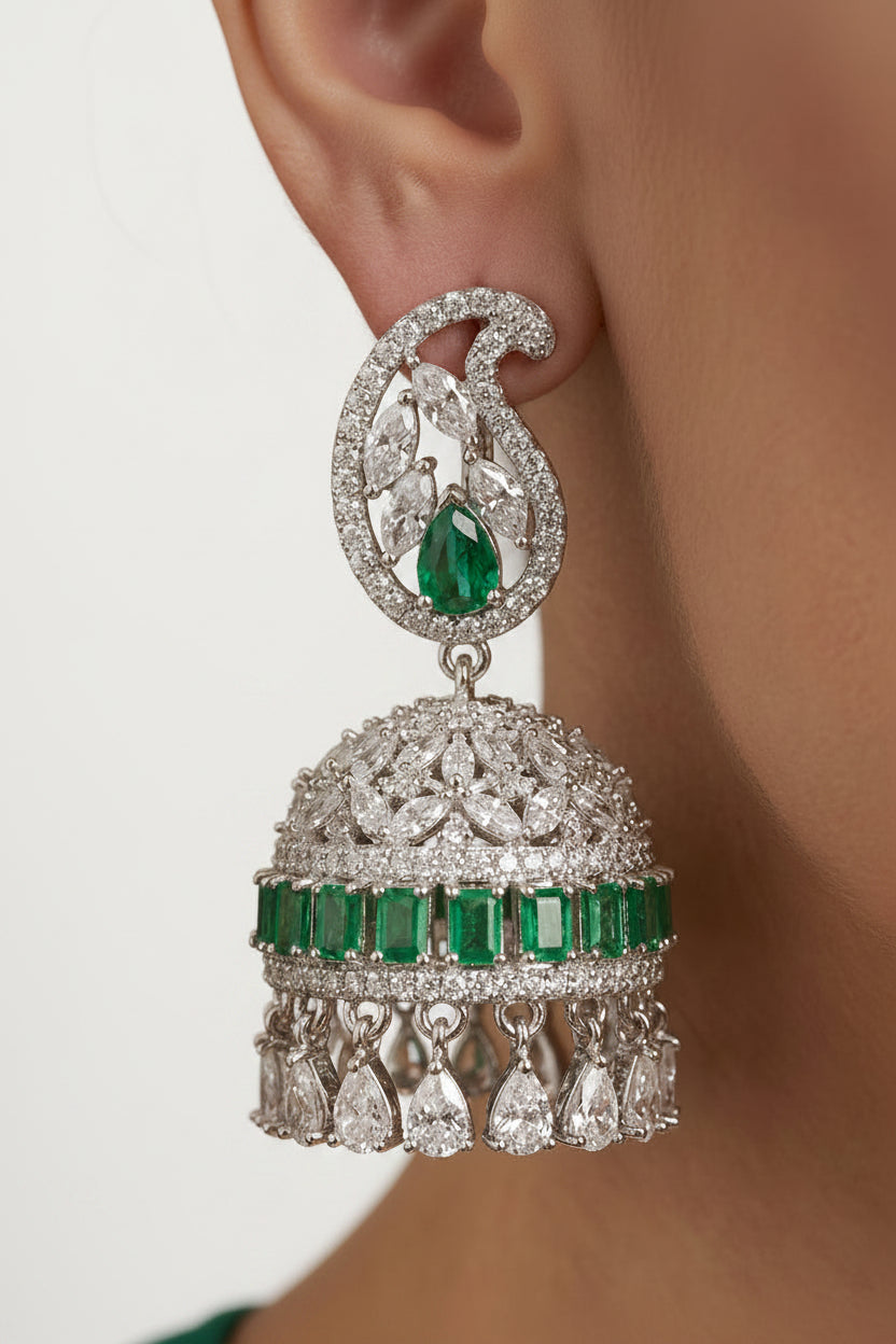 A close-up of a person wearing a large traditional Indian jhumka earring. The earring features a silver-toned paisley-shaped top set with clear stones and a central green emerald-cut gemstone, hanging from a matching dome-shaped lower section decorated with a row of green emerald-cut stones and teardrop dangling crystals.