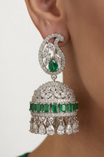 A close-up of a person wearing a large traditional Indian jhumka earring. The earring features a silver-toned paisley-shaped top set with clear stones and a central green emerald-cut gemstone, hanging from a matching dome-shaped lower section decorated with a row of green emerald-cut stones and teardrop dangling crystals.