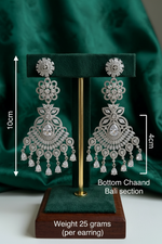 Elegant silver American Diamond chandelier earrings on a green display stand, featuring a floral tops section, intricate leaf motifs, and a detailed 4cm bottom Chaand Bali crescent base with hanging pear-cut zirconia drops.