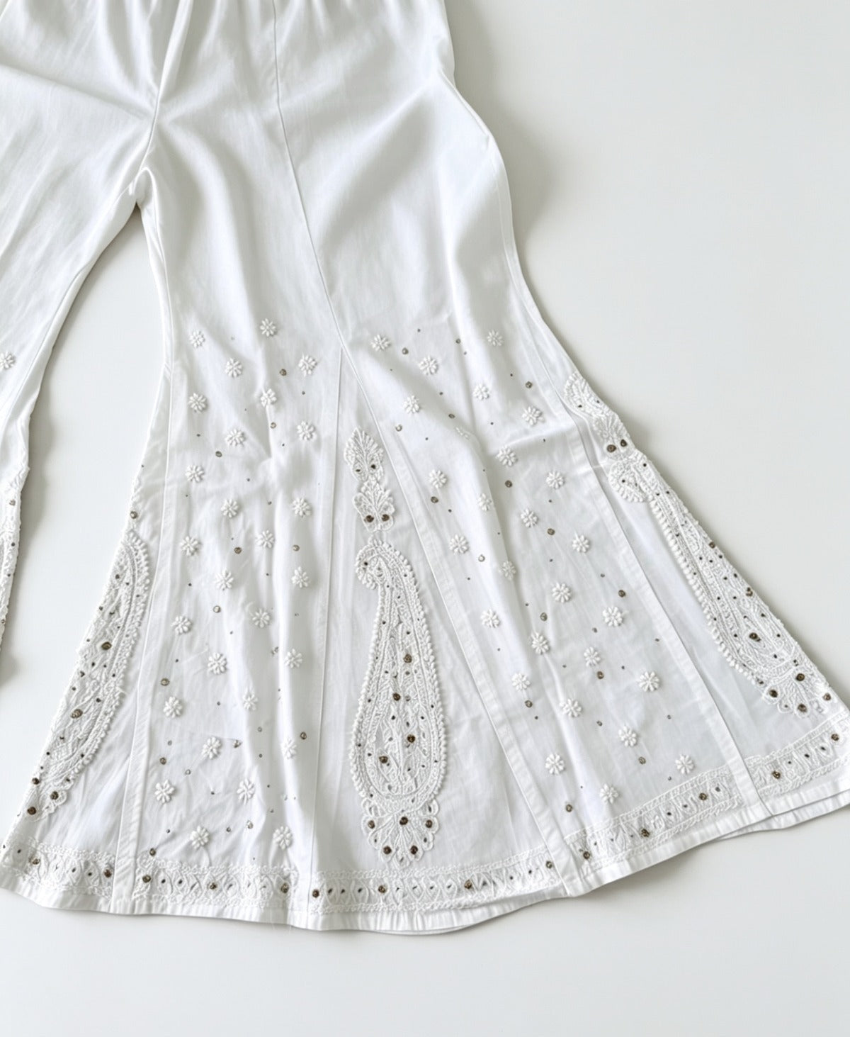 Close-up flat lay of an ivory white sharara featuring detailed floral Bakhiya embroidery, intricate paisley motifs, and subtle metallic accents on the fabric.