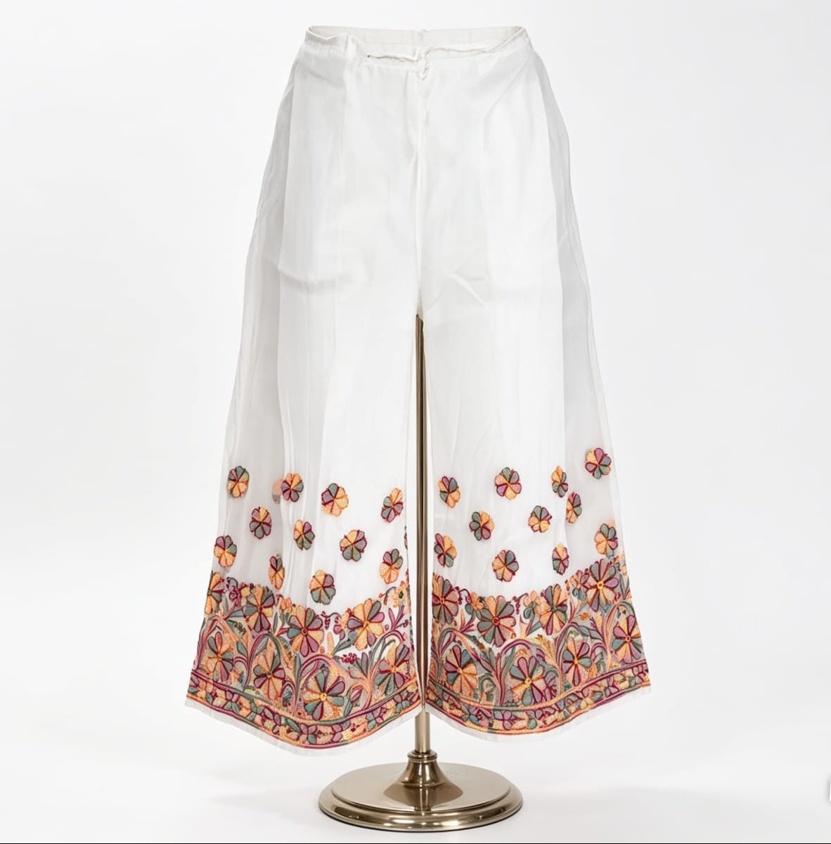 Wide-leg white pants on a mannequin, featuring intricate multi-color floral embroidery at the hem and scattered motifs above. Against a plain white background.