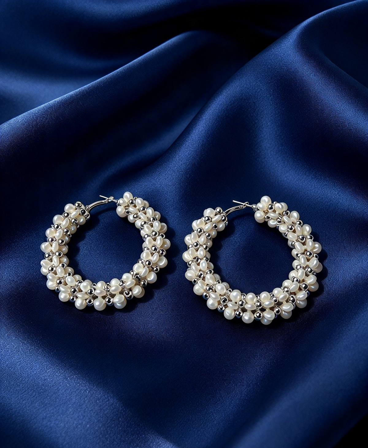 Pair of hand‑woven ivory pearl cluster hoop earrings with polished silver‑tone accents resting on deep navy blue silk satin — Famaza Couture bridal luxury jewelry.