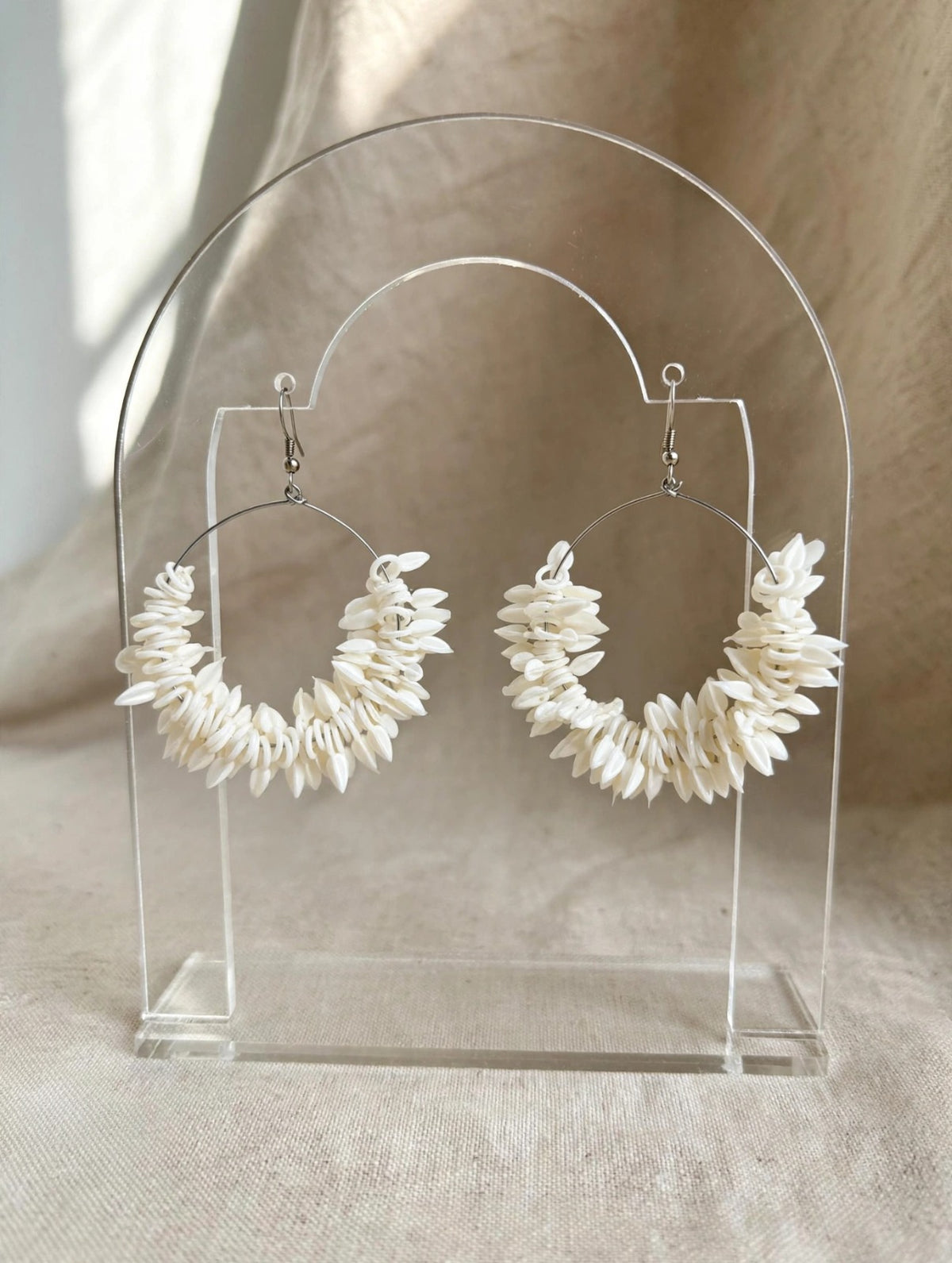 Pair of ivory white natural shell petal cluster hoop earrings on clear acrylic stand, neutral beige linen background — Famaza Couture.