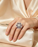 Silver-tone floral halo ring with lavender purple stones and sparkling AD center, worn on hand resting on cream satin clutch with soft silk background