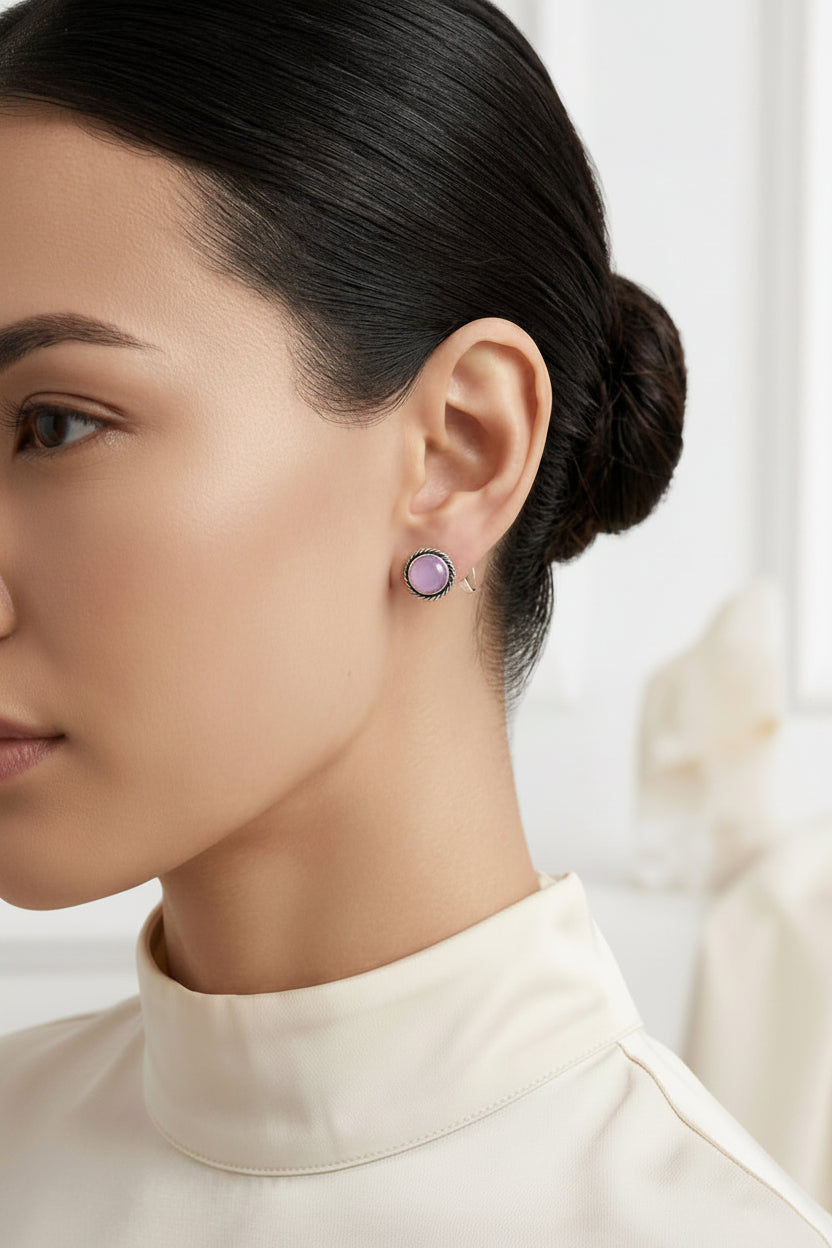 A close-up side profile of a woman with dark hair in a bun wearing a small round lavender purple stud earring, paired with a cream mock neck top.