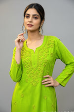 Front view of a woman wearing a vibrant lime green cotton short kurti featuring intricate matching floral embroidery, a keyhole neckline, and a square hem line, styled with traditional earrings while posing with her hand near her face.