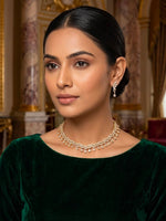 A woman with dark hair pulled back wears a gold-toned choker necklace and matching drop earrings, photographed indoors against a warm ornate background. She is also wearing a dark green velvet top.