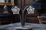A pair of diamond earrings displayed on a black velvet stand in an elegant, dark, luxurious room. The earrings feature round diamond clusters set in silver-toned metal.