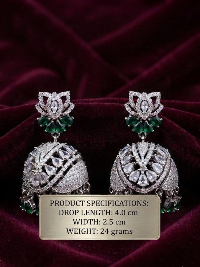 A pair of ornate silver-toned jhumka earrings with green stone accents and clear pavé-style detailing, displayed on a dark burgundy velvet background with a beige product specification label overlaid near the bottom.