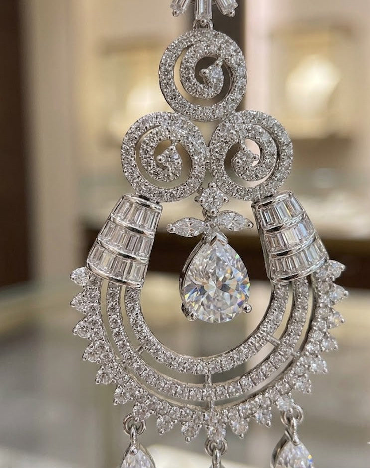 A close-up view of an intricate, silver-toned chandelier earring featuring a central pear-cut crystal, surrounded by detailed filigree metalwork and rows of sparkling baguette and pavé-set stones."