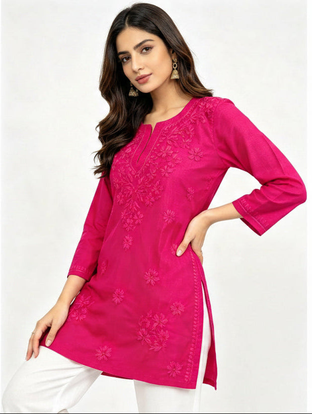 A woman modeling a bright pink embroidered short kurti with white pants against a light background.