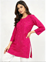 A woman modeling a bright pink embroidered short kurti with white pants against a light background.